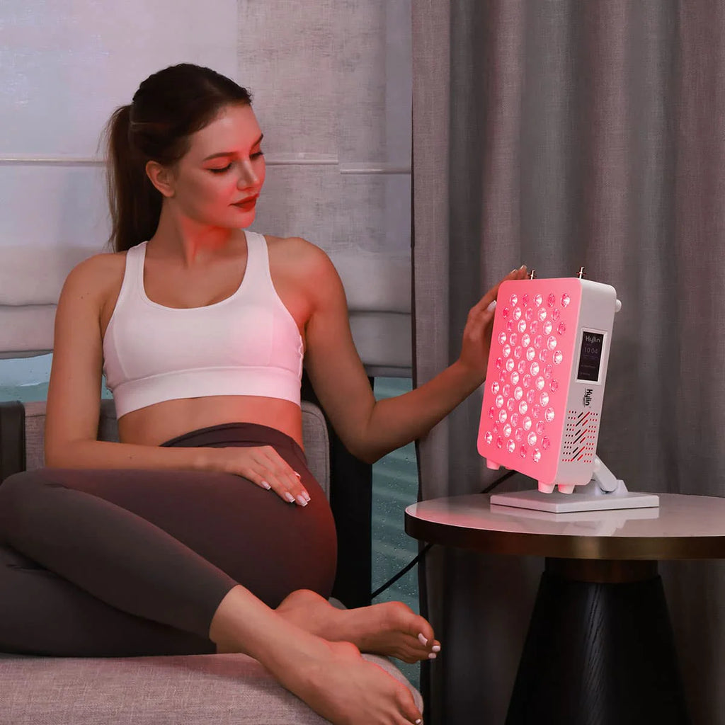 Woman using red LED light therapy panel on sofa couch.